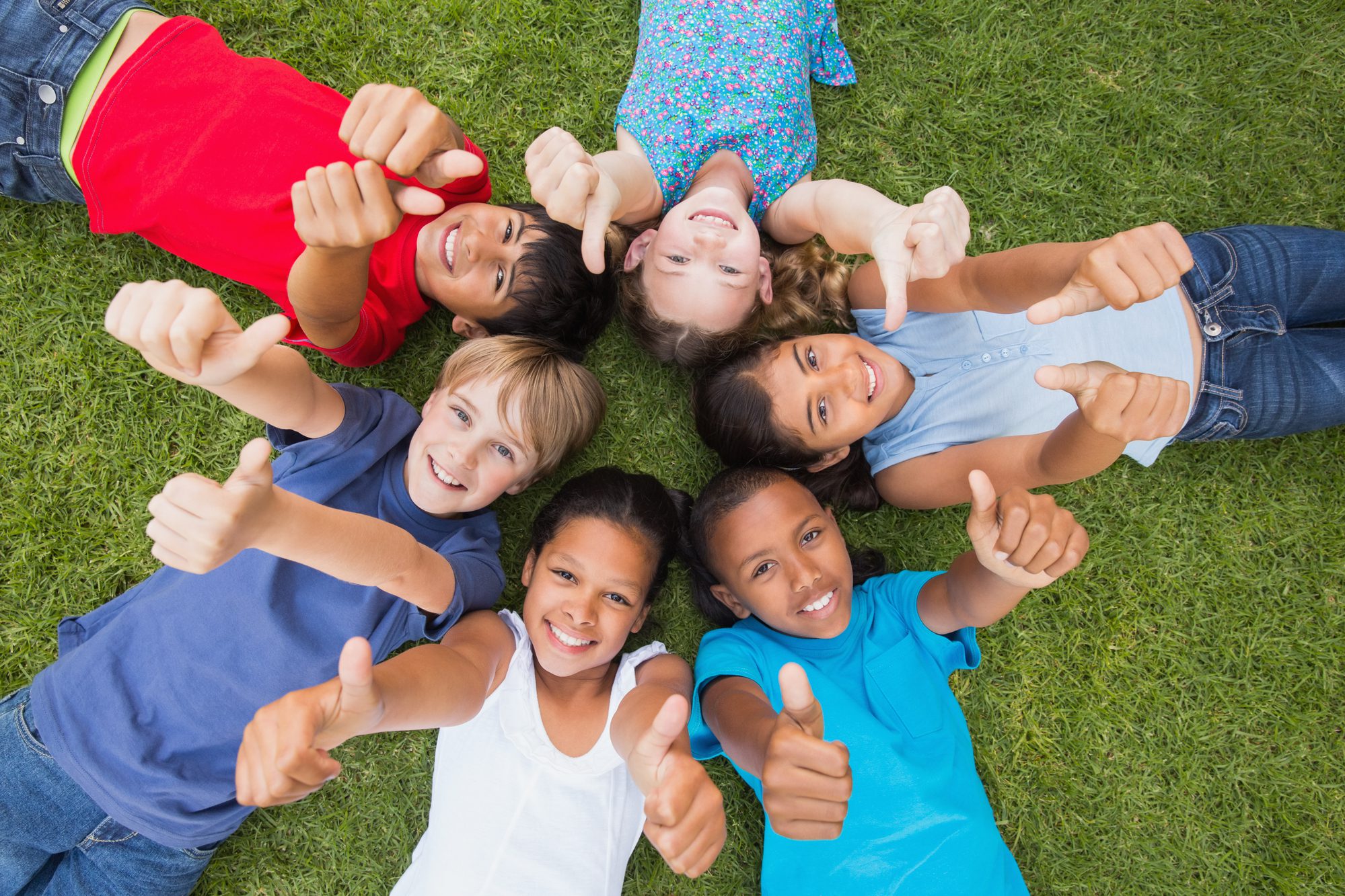 A group of children laying in the grass with thumbs up.